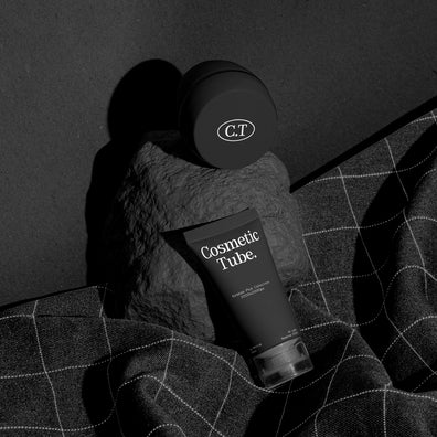 A cosmetic product arrangement featuring a black jar mockup and a black tube mockup labeled 'Cosmetic Tube.' resting on a dark stone against a dark background with draped dark fabric.