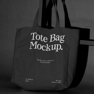 A dark tote bag with large white text reading 'Tote Bag Mockup.' hanging against a dark gray wall, with smaller mockup text below.