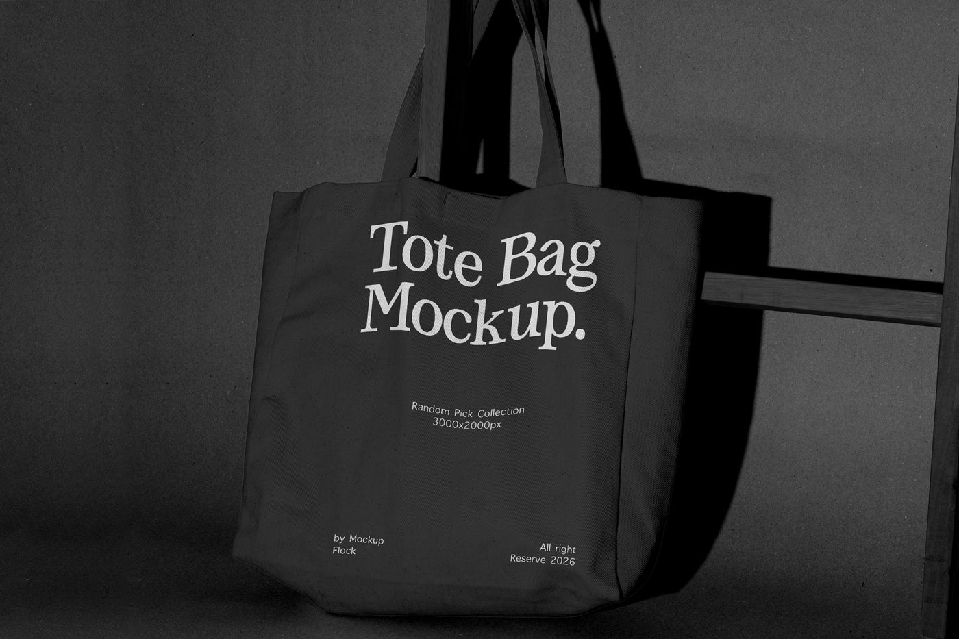 A dark tote bag with large white text reading 'Tote Bag Mockup.' hanging against a dark gray wall, with smaller mockup text below.