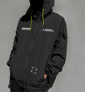 A person wearing a black hooded windbreaker jacket mockup with white printed graphics and bright yellow drawstrings, standing against a textured gray wall.