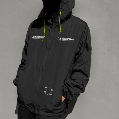 A person wearing a black hooded windbreaker jacket mockup with white printed graphics and bright yellow drawstrings, standing against a textured gray wall.