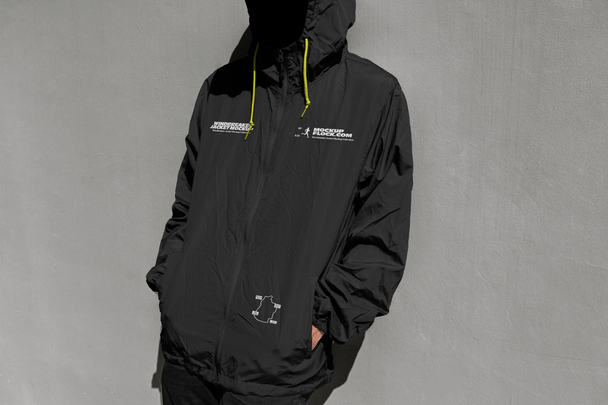 A person wearing a black hooded windbreaker jacket mockup with white printed graphics and bright yellow drawstrings, standing against a textured gray wall.