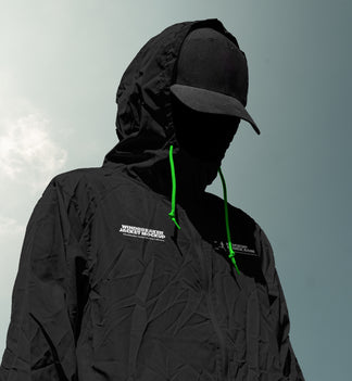 A person wearing a black hooded jacket mockup with bright green drawstrings and a black cap, shown against a pale sky background. The jacket has a small white logo on the chest.
