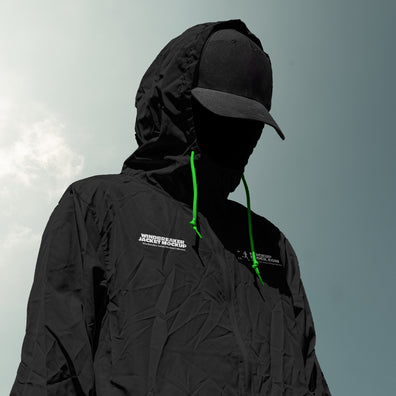 A person wearing a black hooded jacket mockup with bright green drawstrings and a black cap, shown against a pale sky background. The jacket has a small white logo on the chest.