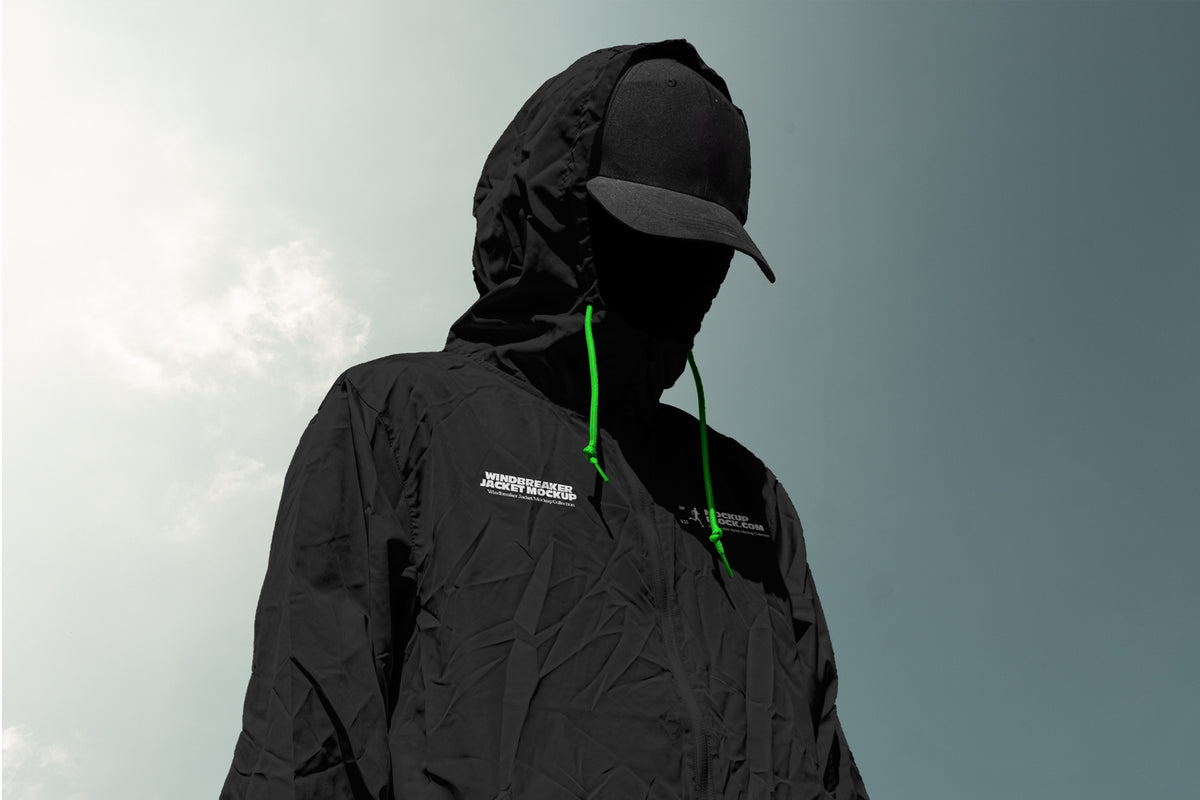 A person wearing a black hooded jacket mockup with bright green drawstrings and a black cap, shown against a pale sky background. The jacket has a small white logo on the chest.