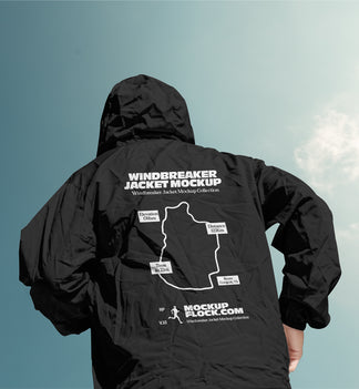 A person viewed from behind wearing a black hooded windbreaker jacket with a large white mockup graphic and text printed on the back, set against a blue sky with light clouds.