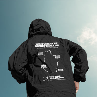 A person viewed from behind wearing a black hooded windbreaker jacket with a large white mockup graphic and text printed on the back, set against a blue sky with light clouds.