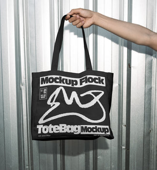 Hand holding a black tote bag featuring an urban graphic design against a silver corrugated metal wall