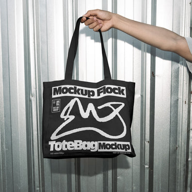 Hand holding a black tote bag featuring an urban graphic design against a silver corrugated metal wall