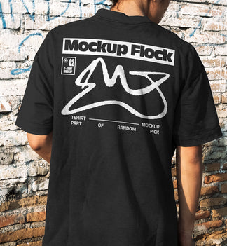 Back view of a person wearing a black streetwear t-shirt with custom graphic design, standing against a distressed brick wall