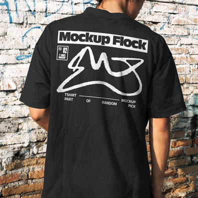 Back view of a person wearing a black streetwear t-shirt with custom graphic design, standing against a distressed brick wall