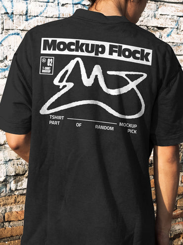 Back view of a person wearing a black streetwear t-shirt with custom graphic design, standing against a distressed brick wall