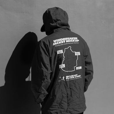 A person viewed from behind wearing a dark hooded jacket mockup with white printed graphics and text on the back, standing against a gray wall with a strong shadow cast to the left.