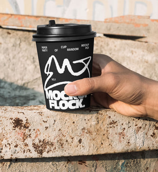 Close-up of a hand holding a black paper coffee cup with a custom logo design against an industrial rusty metal background