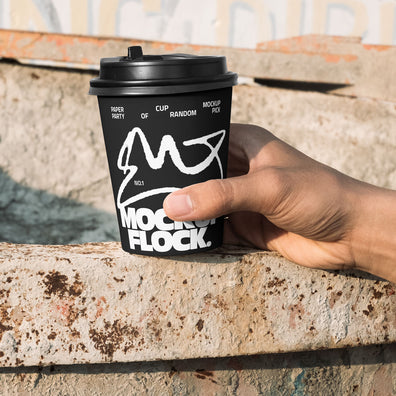 Close-up of a hand holding a black paper coffee cup with a custom logo design against an industrial rusty metal background