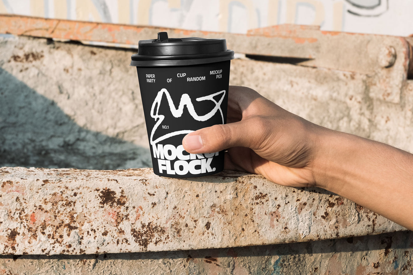 Close-up of a hand holding a black paper coffee cup with a custom logo design against an industrial rusty metal background
