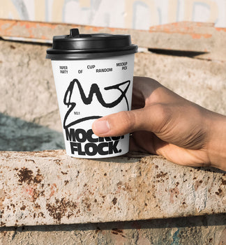 Close-up of a hand holding a white paper coffee cup with a custom logo design against an industrial rusty metal background