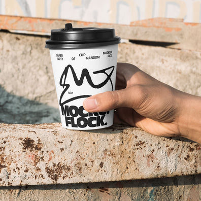 Close-up of a hand holding a white paper coffee cup with a custom logo design against an industrial rusty metal background