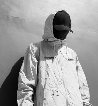 A person wearing a light-colored hooded jacket mockup, a black baseball cap, and a black face covering stands against a textured gray wall, casting a dark shadow.