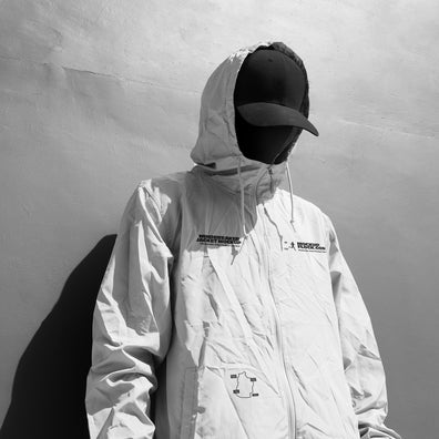 A person wearing a light-colored hooded jacket mockup, a black baseball cap, and a black face covering stands against a textured gray wall, casting a dark shadow.