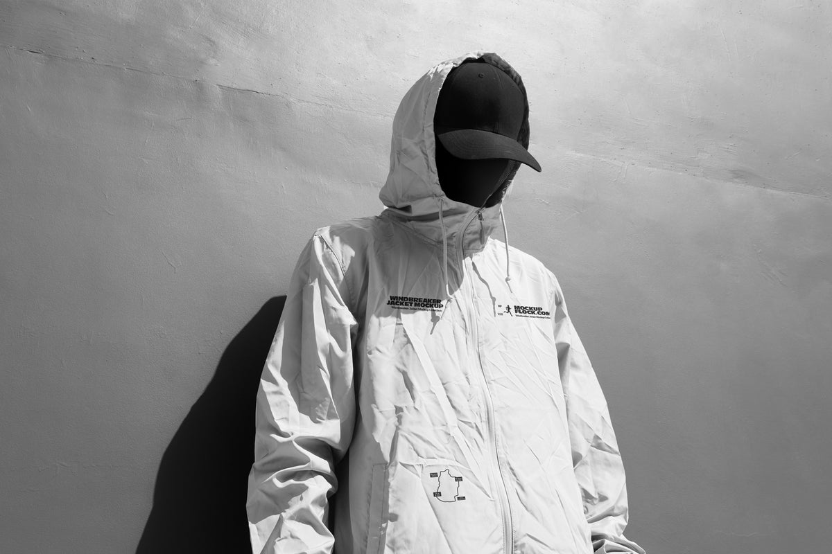 A person wearing a light-colored hooded jacket mockup, a black baseball cap, and a black face covering stands against a textured gray wall, casting a dark shadow.