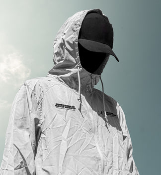 A person wearing a light gray hooded windbreaker jacket mockup with black logo text on the chest, a black baseball cap, and a black face covering, photographed outdoors against a pale sky.