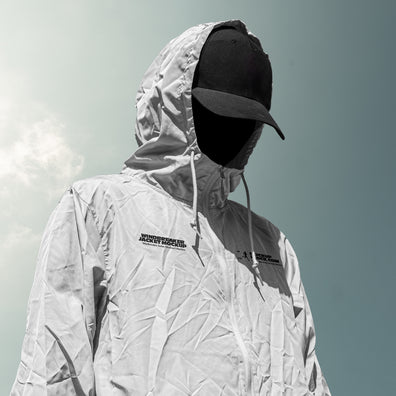 A person wearing a light gray hooded windbreaker jacket mockup with black logo text on the chest, a black baseball cap, and a black face covering, photographed outdoors against a pale sky.