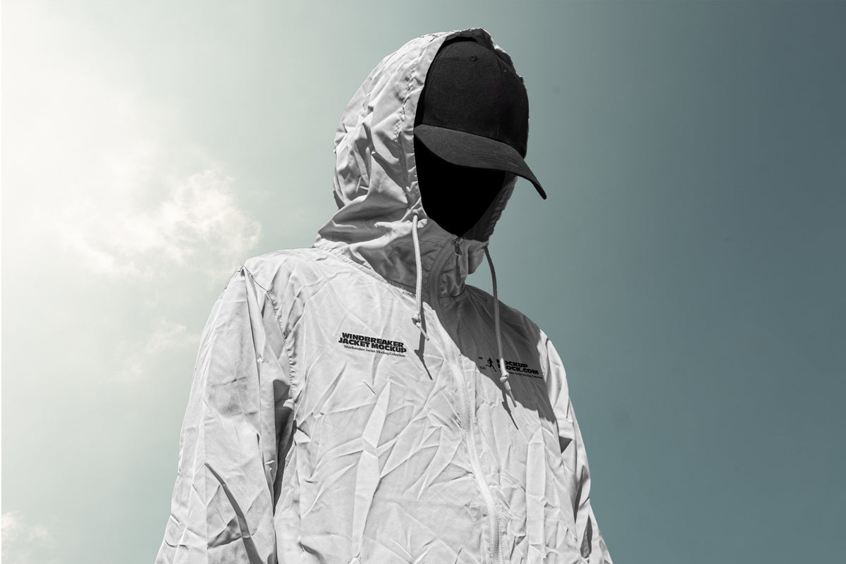 A person wearing a light gray hooded windbreaker jacket mockup with black logo text on the chest, a black baseball cap, and a black face covering, photographed outdoors against a pale sky.