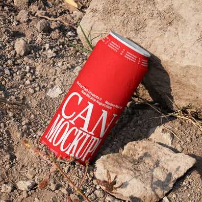 Red beverage can leaning against a rock on dry dirt ground with dried grass, outdoor mockup