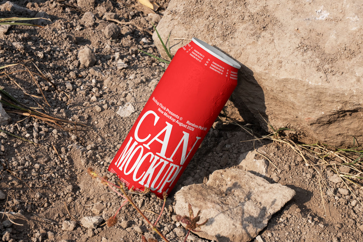 Red beverage can leaning against a rock on dry dirt ground with dried grass, outdoor mockup