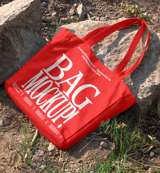 Red canvas tote bag mockup with long handles lying on rocks and dirt ground outdoors, mockup