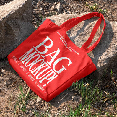 Red canvas tote bag mockup with long handles lying on rocks and dirt ground outdoors, mockup