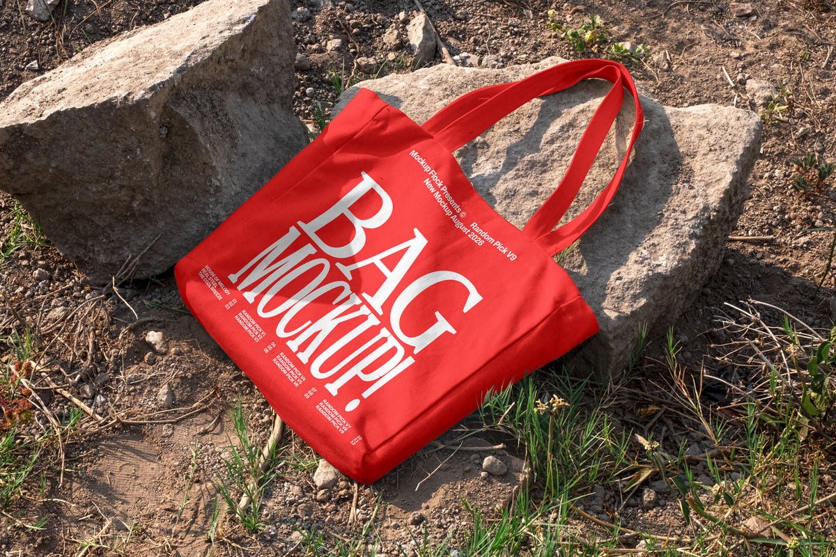 Red canvas tote bag mockup with long handles lying on rocks and dirt ground outdoors, mockup