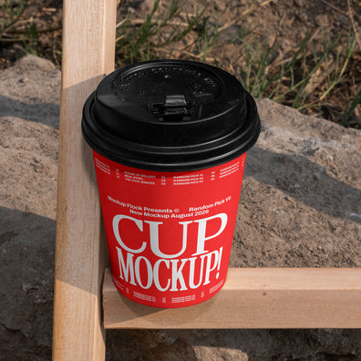 Red paper coffee cup mockup with black lid placed on a wooden fence beam with rocky dirt background outdoors