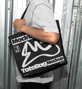 Person carrying a black canvas tote bag over the shoulder with custom graphic print, standing in front of an industrial metal door
