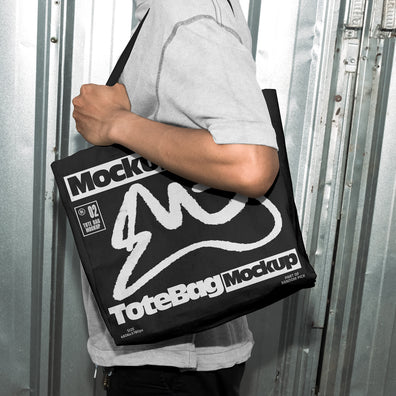 Person carrying a black canvas tote bag over the shoulder with custom graphic print, standing in front of an industrial metal door