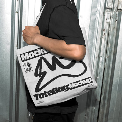 Person carrying a white canvas tote bag over the shoulder with custom graphic print, standing in front of an industrial metal door