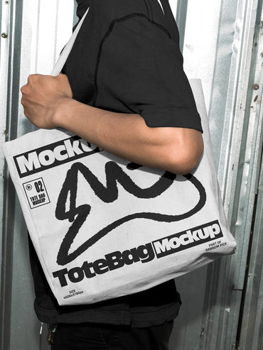 Person carrying a white canvas tote bag over the shoulder with custom graphic print, standing in front of an industrial metal door