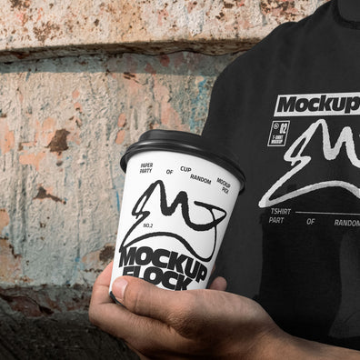 Person wearing a black t-shirt holding a white paper coffee cup with custom branding against an urban textured wall