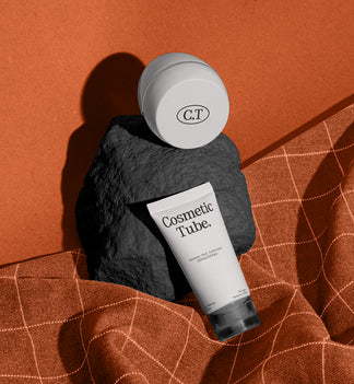 A cosmetic product arrangement featuring a white jar mockup and a white tube mockup labeled 'Cosmetic Tube.' resting on a dark stone against a warm terracotta background with draped orange fabric.