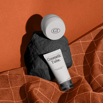 A cosmetic product arrangement featuring a white jar mockup and a white tube mockup labeled 'Cosmetic Tube.' resting on a dark stone against a warm terracotta background with draped orange fabric.