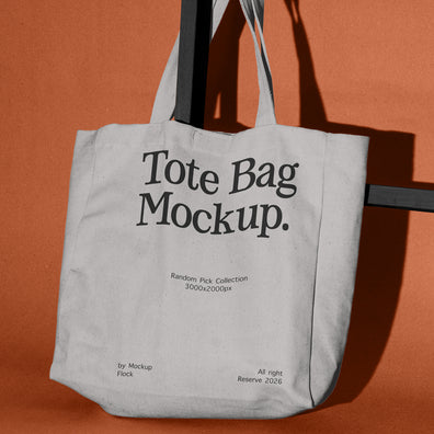 A light gray canvas tote bag hanging from a black metal stand against a solid terracotta orange background. The bag features placeholder black text reading 'Tote Bag Mockup.