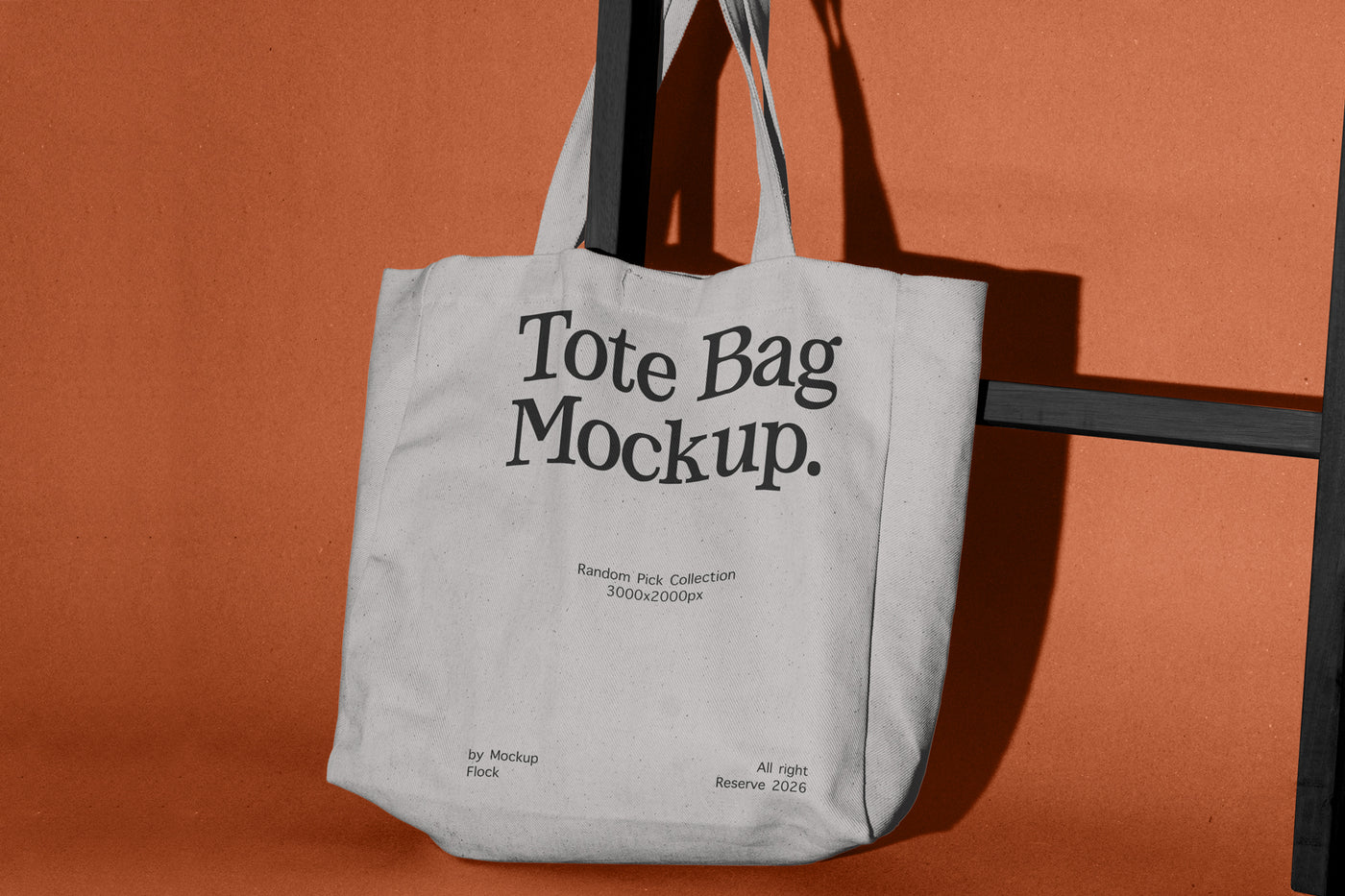A light gray canvas tote bag hanging from a black metal stand against a solid terracotta orange background. The bag features placeholder black text reading 'Tote Bag Mockup.