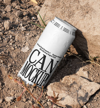 White beverage can leaning against a rock on dry dirt ground with dried grass, outdoor mockup