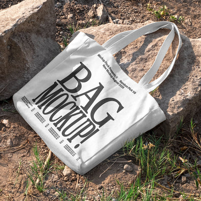 White canvas tote bag mockup with long handles lying on rocks and dirt ground outdoors, mockup