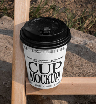 White paper coffee cup mockup with black lid placed on a wooden fence beam with rocky dirt background outdoors