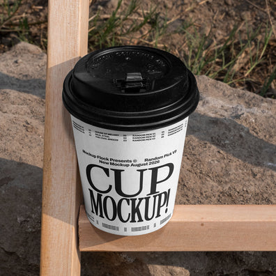 White paper coffee cup mockup with black lid placed on a wooden fence beam with rocky dirt background outdoors
