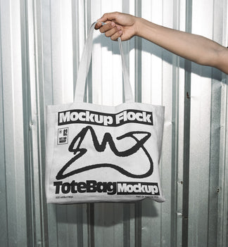 Hand holding a white tote bag featuring an urban graphic design against a silver corrugated metal wall