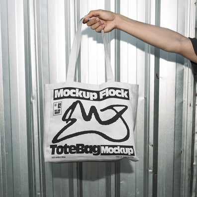 Hand holding a white tote bag featuring an urban graphic design against a silver corrugated metal wall