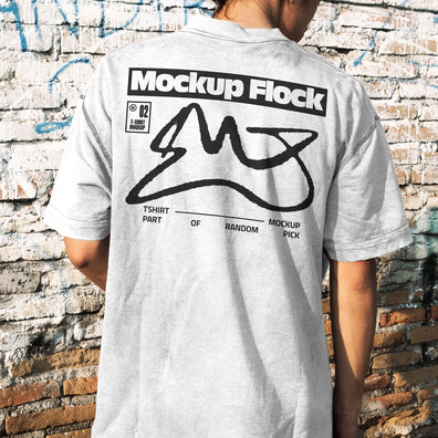 Back view of a person wearing a white streetwear t-shirt with custom graphic design, standing against a distressed brick wall